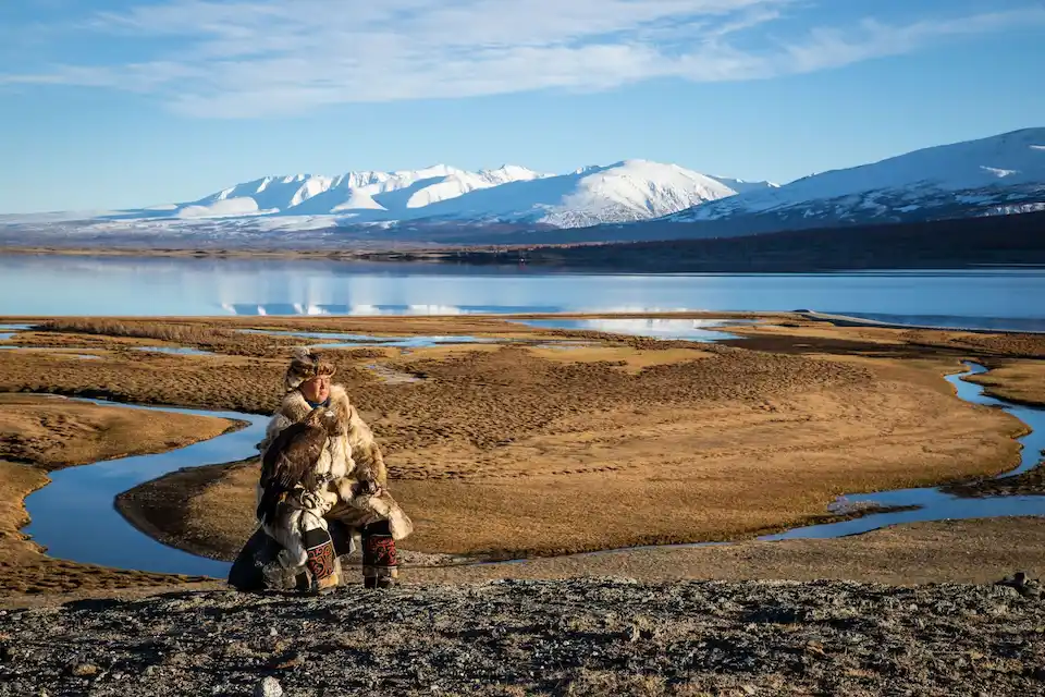 Mongolian eagle hunter in traditional fur clothing sitting on a rock in western Mongolia with his golden eagle, overlooking vast steppe, river, and snow‑capped Altai Mountains.