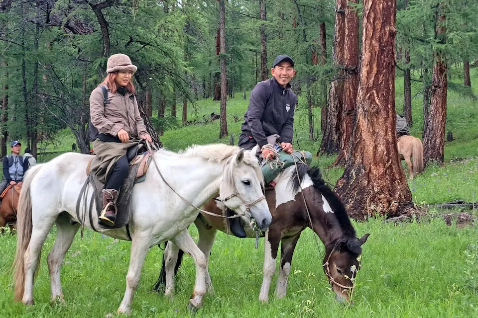 Riders journey through a lush Mongolian forest on white horses, accompanied by a grazing foal and surrounded by tall trees and vibrant greenery.