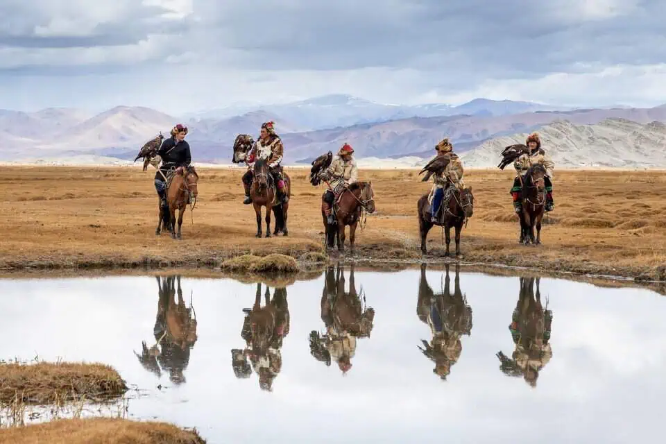 Five eagle hunters ride across the steppe, their golden eagles and traditional attire mirrored in the still water. A rare glimpse into Mongolia’s living heritage.